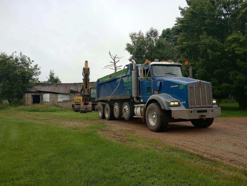 Blue dump truck on gravel road near a construction site and old barn in a rural area.