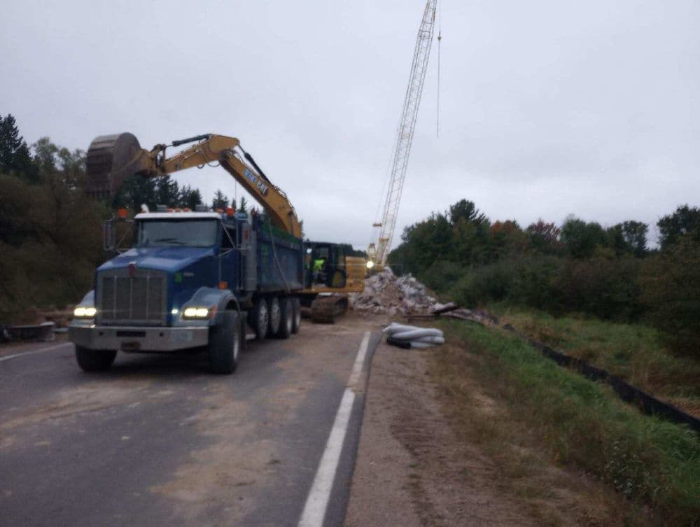 Construction site with heavy machinery, including a dump truck and crane, on a rural road.