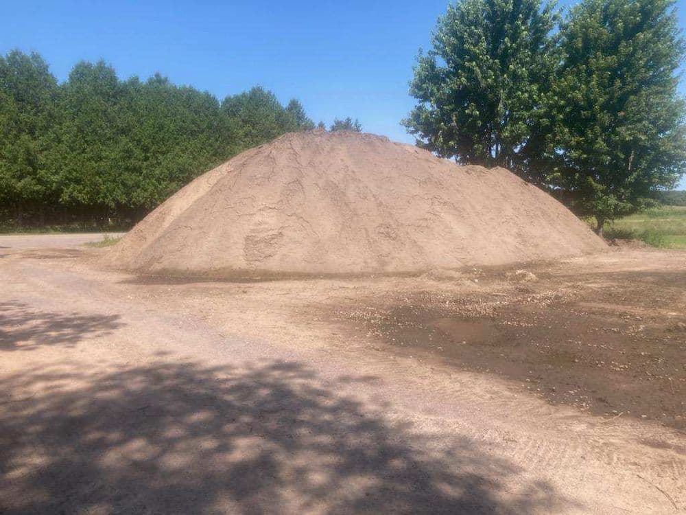 Large sand pile on dirt lot with trees in background under clear blue sky.